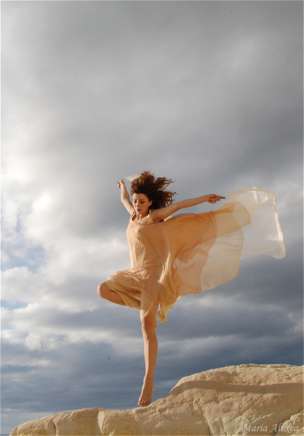 Maria Alexea in a pale flowing dress poised on a rock beneath a dramatic sky