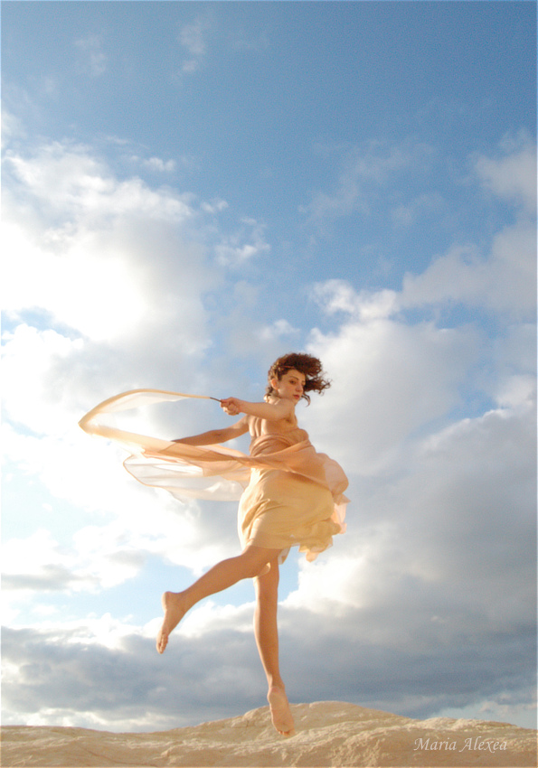 Maria Alexea in a pale flowing dress moving across a rocky landscape beneath a bright sky