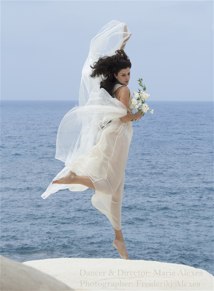 Maria Alexea in a flowing white dress, leaping above the sea while holding a bouquet of white roses.